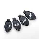 Four Fourteen Golf black shield-shaped interchangeable club number tags numbered 3, 4, 5, and X with white embroidered numerals and nylon pull loops, displayed on white background