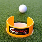 A yellow golf putting cup with a hole, designed to aid in practicing putting, positioned on a green surface with a white golf ball in the background.