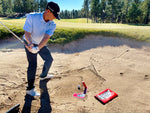 Golfer mid-downswing approaching the EyeLine Golf Bunker Pro System board and entry point peg in a course bunker during a sand shot drill