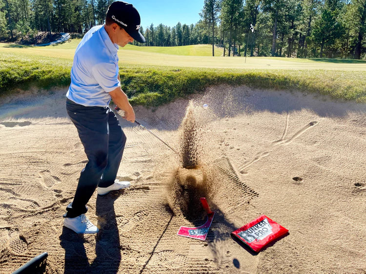 Golfer executing a bunker shot with the EyeLine Golf Bunker Pro System board and sand wedge, ball launching out of the sand with an explosion of sand visible
