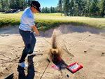 Golfer executing a bunker shot with the EyeLine Golf Bunker Pro System board and sand wedge, ball launching out of the sand with an explosion of sand visible