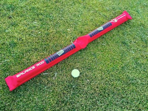 EyeLine Golf Balance Rod laid flat on grass beside a golf ball, showing the full length of the inflatable red balance training aid