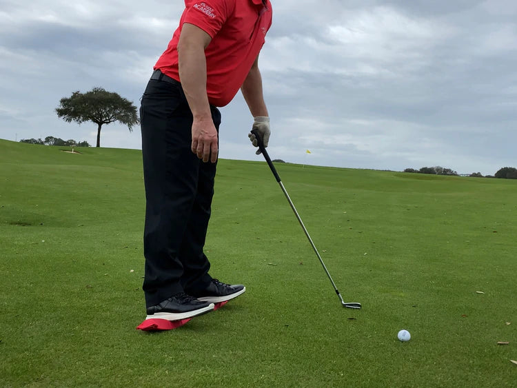 Golfer in address position with a wedge, red EyeLine Golf Balance Rod placed under both feet to train lower body stability during a chip shot