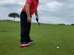 Golfer in address position with a wedge, red EyeLine Golf Balance Rod placed under both feet to train lower body stability during a chip shot