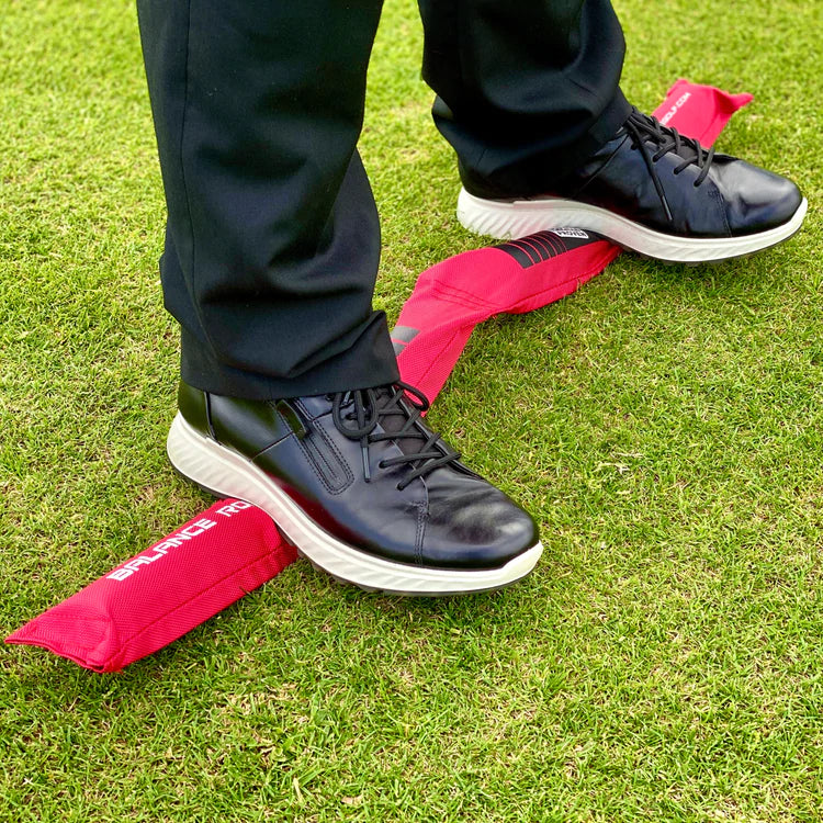 A person standing on a red balance rod with black shoes and trousers on, positioned on a green grass surface.