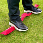 A person standing on a red balance rod with black shoes and trousers on, positioned on a green grass surface.