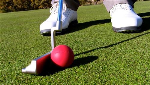 Ground-level view of a putter making contact with the EyeLine Golf Ball of Steel red weighted ball on a practice green