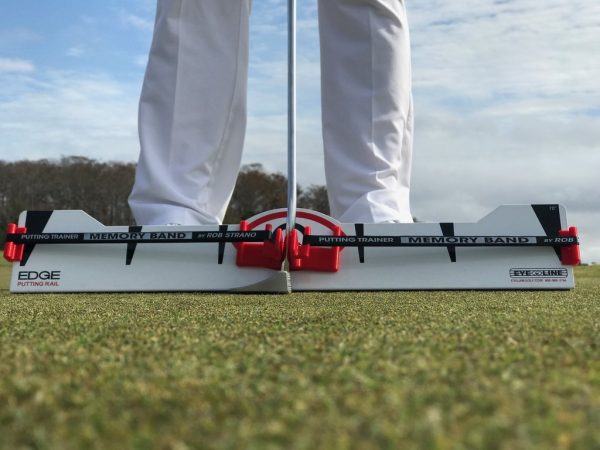 Front-on ground-level view of golfer's stance over the EyeLine Golf Edge Putting Rail with Memory Band system, showing the red arc trainer and foot alignment guides on either side