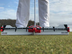 Front-on ground-level view of golfer's stance over the EyeLine Golf Edge Putting Rail with Memory Band system, showing the red arc trainer and foot alignment guides on either side