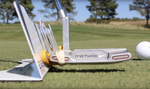 Ground-level view of the EyeLine Golf Putter Guide attached to a blade putter face beside the Edge training aid and a golf ball on a course green