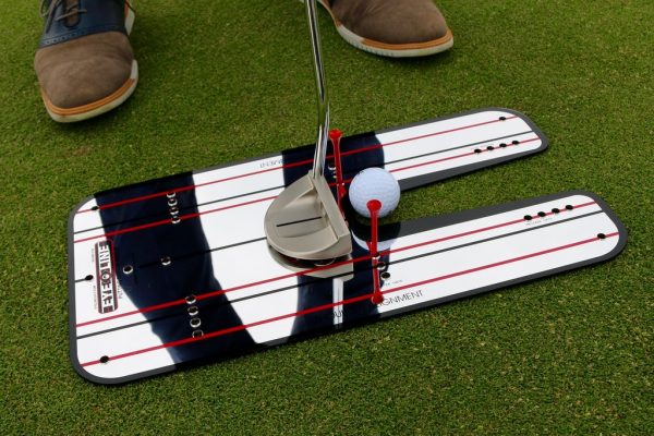 Angled view of putter and golf ball at address on the EyeLine Golf Classic Putting Mirror with red tee gate, showing the U-shaped putter channel and red alignment lines reflected beneath the player's stance