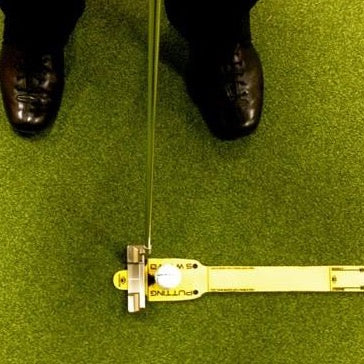 A close-up of a golf putting sword with a yellow and black color scheme, positioned on a green mat with a person's feet in black shoes in the background.