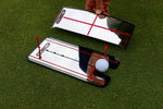 EyeLine Golf putting alignment mirror with red tee gate setup alongside a smaller travel mirror, golf ball positioned at the putter gate on the green