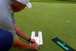 Golfer positioning the EyeLine Golf Spot On Laser alignment board on the green, with the laser target visible in the distance and the Spot On carry pouch beside it