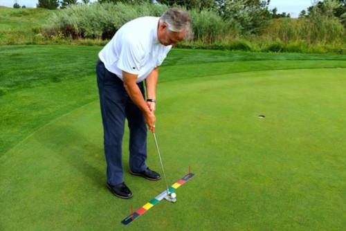 Golfer putting on a course green with the EyeLine Golf Stroke Meter laid flat, colour-coded zones visible from the side as he addresses the ball toward the hole