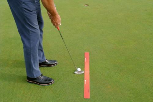 Golfer addressing a putt with a red alignment strip placed on the green to guide putter path and ball position toward the hole