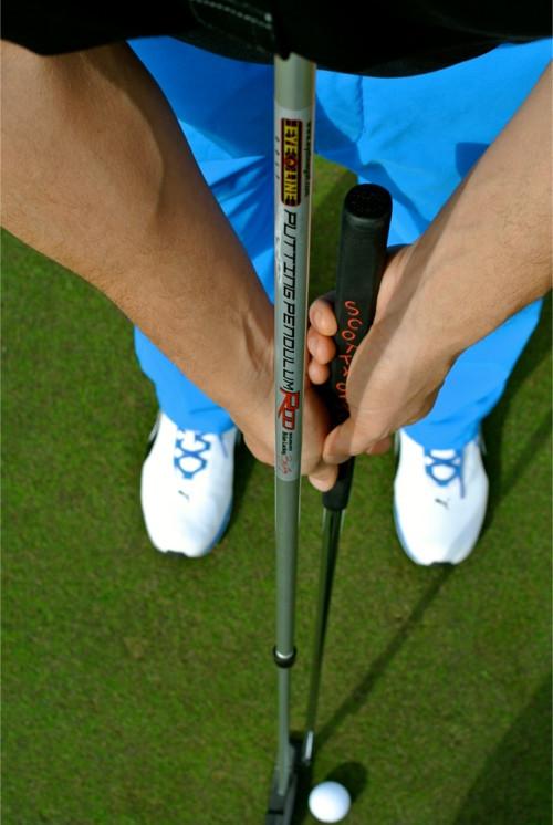 Overhead view of golfer gripping the Eyeline Golf Putting Pendulum Rod alongside a putter at address over a golf ball on the green