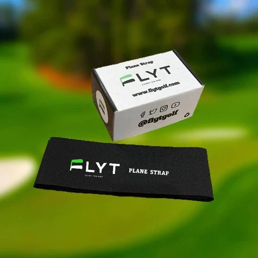 A black golf plane strap with the FLYT brand logo, displayed in front of a blurred background featuring a golf course. A white box with the FLYT logo is also visible in the background.